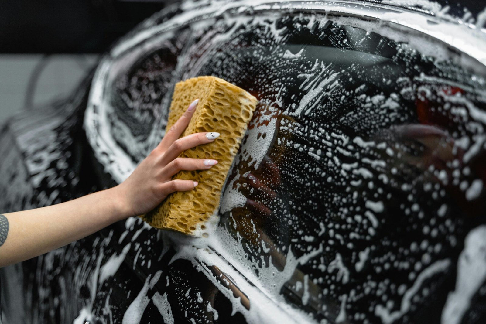 A hand scrubs a car window with a sponge, covered in soap and foam, at a car wash.